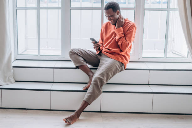 Cheerful black man with smartphone sits on large window sill stock photo