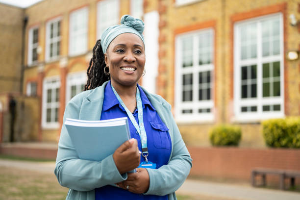 Cheerful Black teacher standing outside education building Candid portrait of smiling early 50s woman in businesswear and headscarf holding stack of composition booklets and looking away from camera. work as a teacher in the uk stock pictures, royalty-free photos & images
