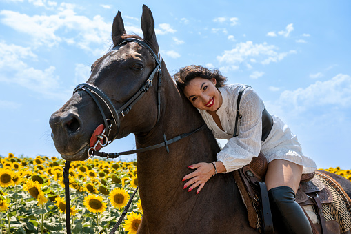 Beautiful brunette riding brown horse. She is smiling at camera. Woman is wearing red lipstick, long black boots and white dress. Sunny day. Sunflower field in background.