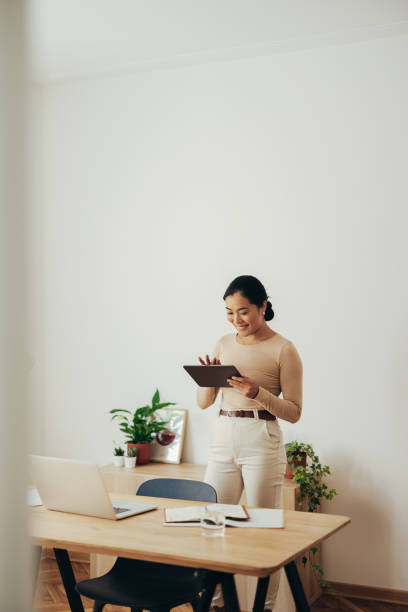 Cheerful Business Woman Using Digital Tablet at Home Happy Asian businesswoman typing something on her digital tablet while standing in a modern office business stock pictures, royalty-free photos & images