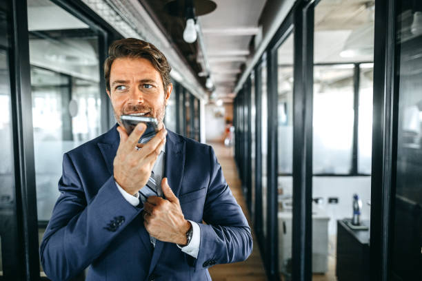 Cheerful businessman using smart phone for sending voice message Happy businessman talking over phone along office corridor, using smart phone for sending voice message speech recognition stock pictures, royalty-free photos & images