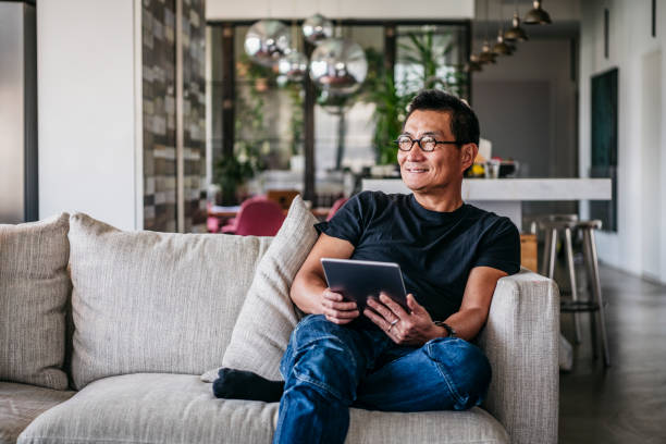 Cheerful Chinese man with tablet looking away and smiling Man in his 50s sitting on sofa at home, leisure time, relaxation, enjoyment using digital tablet stock pictures, royalty-free photos & images