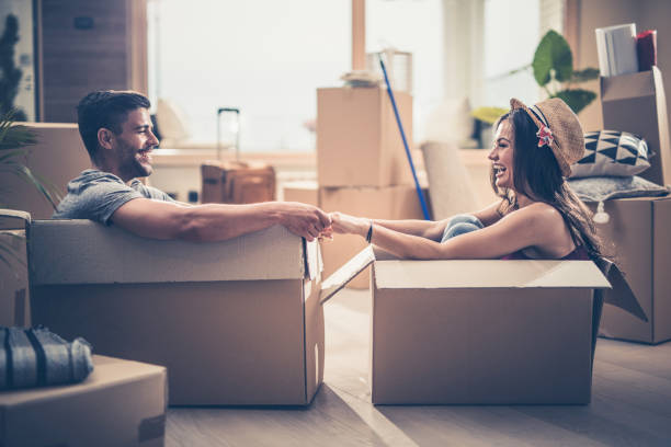 Cheerful couple having fun while sitting in cardboard boxes at their new home. Young happy couple holding hands and having fun while sitting in cardboard boxes at their new apartment. flat physical description stock pictures, royalty-free photos & images