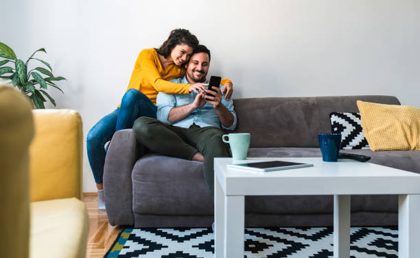 Cheerful couple using mobile phone stock photo Man holding in hands phone and woman showing screen and smiling together embraced while sitting on sofa at home couple relationship stock pictures, royalty-free photos & images