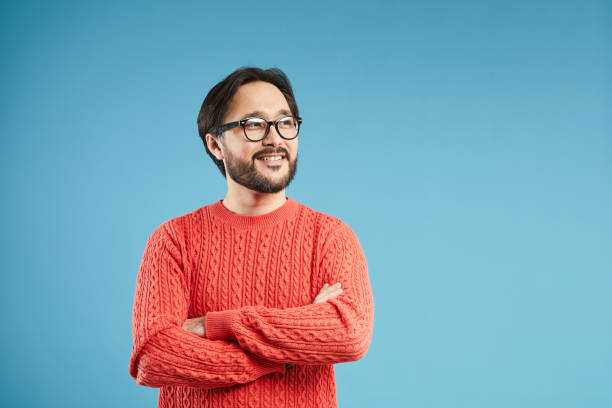 vrolijke dromerige jonge aziatische man met baard en snor dragen stijlvolle trui staande tegen blauwe achtergrond en kijken weg terwijl ze dromerige - gekleurde achtergrond stockfoto's en -beelden