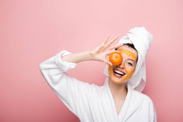cheerful girl with citrus facial mask covering eye with tangerine on pink background cheerful girl with citrus facial mask covering eye with tangerine on pink background orange face mask stock pictures, royalty-free photos & images