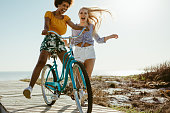 Cheerful female friends having fun with a cycle outdoors. Two girls playing with a bicycle on the boardwalk on seaside.
