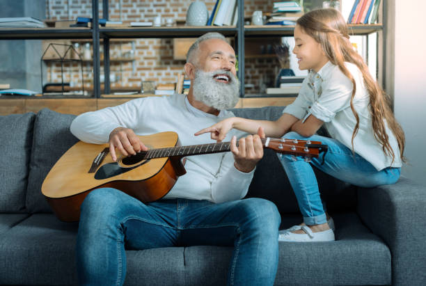 Cheerful grandfather singing for his adorable granddaughter Her first serenade. Beautiful young lady smiling while sitting on a sofa and listening to her talented granddad singing and playing guitar. baby boomers stock pictures, royalty-free photos & images