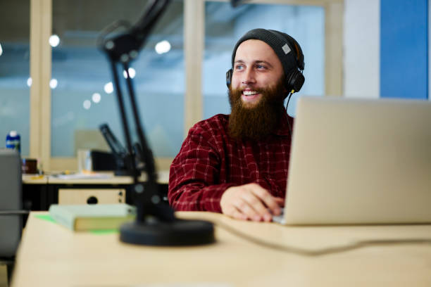 Cheerful graphic designer enjoying favorite compositions playing over cool headphones during work break.Bearded smiling copywriter listening good music while looking away sitting in office stock photo