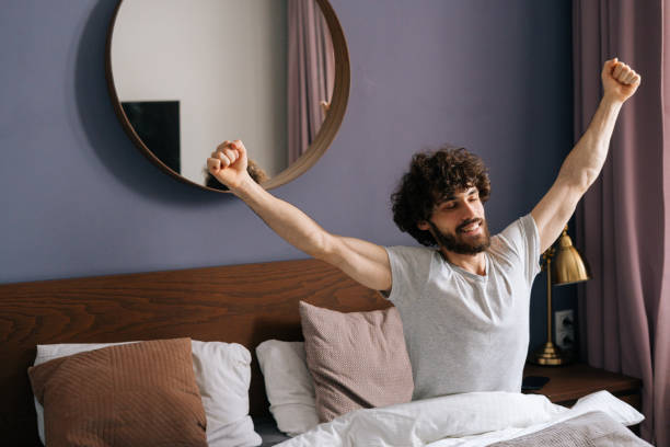 Cheerful handsome young man waking up and stretching arms in morning sitting on bed. Happy bearded male have pleasant dream. stock photo