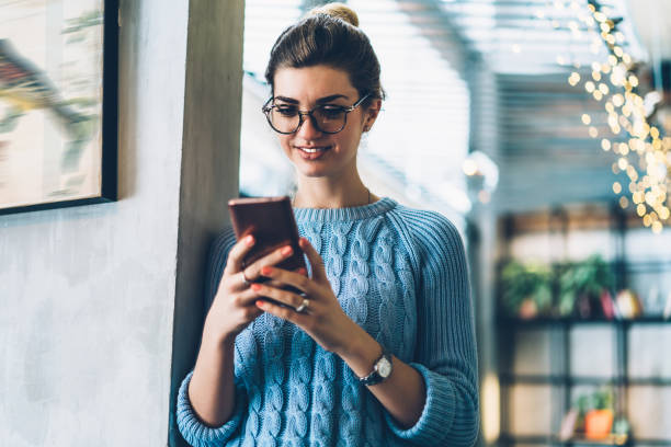 Cheerful hipster blogger in eyeglasses reading funny comments under internet post holding modern smartphone in hands.Positive young woman laughing during online chat with friends on cellular stock photo