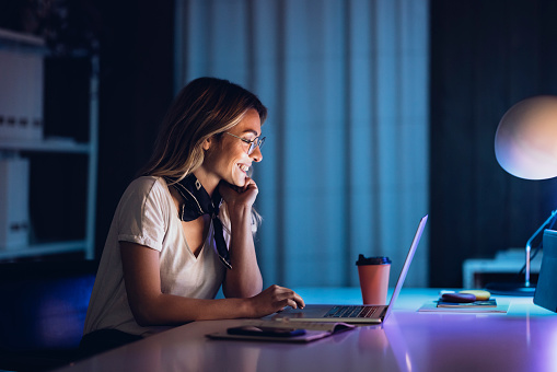 Side view of smiling blond woman in glasses sitting at office table in night and watching laptop having cup of coffee aside