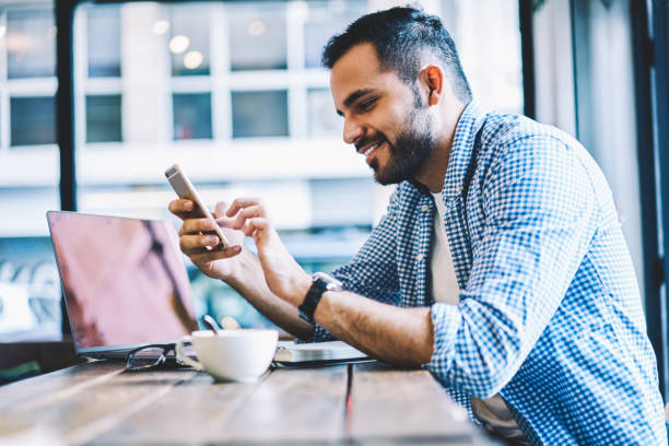 Cheerful male person text messages on smartphone chatting in social networks during coffee break,smiling young man checking mail on cellular pleased with getting discount for mobile internet stock photo