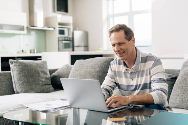 fröhlicher mann lächelnd, während mit laptop in der nähe von kreditkarte im wohnzimmer - einzelner mann über 40 stock-fotos und bilder
