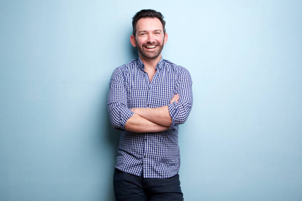cheerful man with beard posing against blue wall with arms crossed stock photo