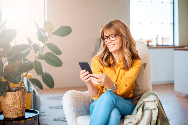 Cheerful mature woman using mobile phone while relaxing in the armchair at home stock photo