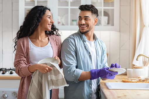 Cheerful Middle Eastern Couple Sharing Domestic Chores, Washing Dishes Together In Kitchen, Happy Millennial Arab Spouses Enjoying Making Cleaning At Home, Looking And Smiling To Each Other