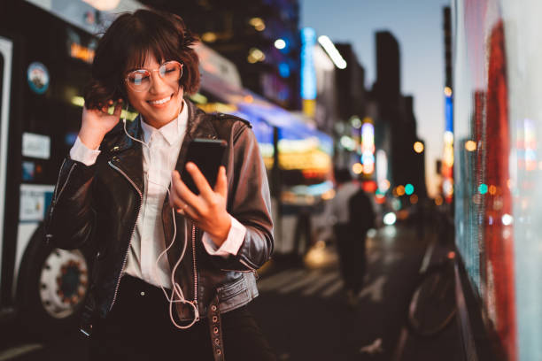 glad söt ung kvinna i coola glasögon och trendiga slitage gå på metropolis street med nattlampor njuter av ljud låtar från spellistan i hörlurar anslutna till smartphone-enhet - hipster person bildbanksfoton och bilder