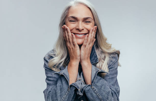 Cheerful senior woman Cheerful senior woman with hands on her face against grey background. Happy mature woman in casual denim shirt. mature women stock pictures, royalty-free photos & images