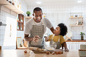 istock Cheerful smiling Black son enjoying playing with his father while doing bakery at home. Playful African family having fun cooking baking cake or cookies in kitchen together. 1311089680
