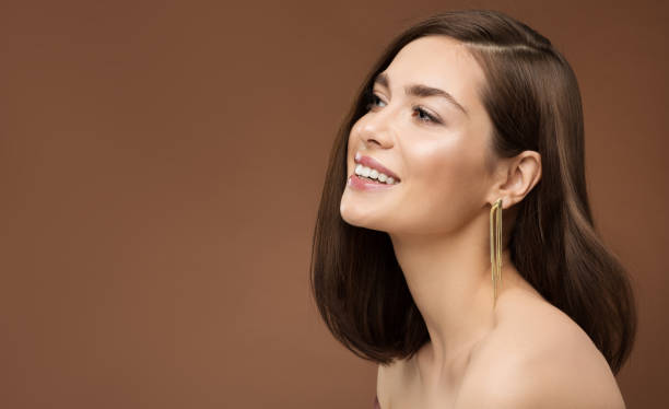 Cheerful Smiling Model Profile Side View. Brunette Beauty Woman with Nude Make up Smooth Skin and Golden Earring over Brown Background stock photo