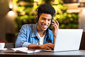 Cheerful black young guy with headset looking at laptop, having fun while studying, cafe interior