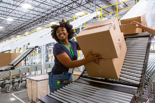 A grinning woman collecting boxes at the bottom of a sorting chute and pushing them along an extendable conveyor belt towards a truck loading dock for delivery.