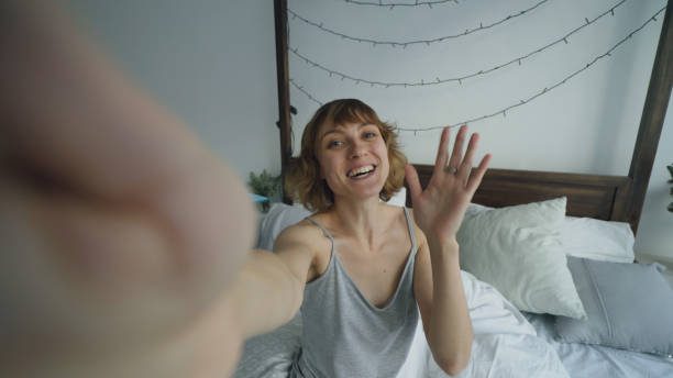 POV of cheerful woman having online video chat with friends using smartphone camera while sitting on bed at home stock photo