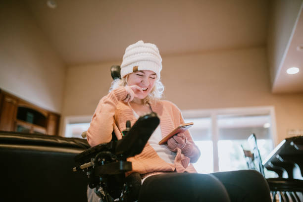 Cheerful Woman In Wheelchair on Smartphone At Home An independent young adult woman with cerebral palsy going about some of her daily routines at home.  She smiles while browsing social media apps on her phone. physical disability stock pictures, royalty-free photos & images