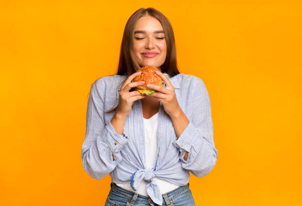 fröhliche frau riecht burger stehend über gelben hintergrund - woman happy eat stock-fotos und bilder