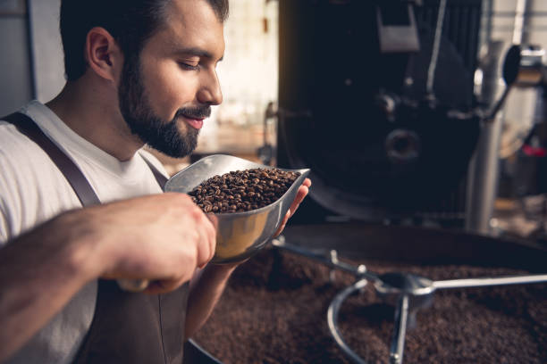 Cheerful worker enjoying beans aroma Side view smiling unshaven master smelling coffee grains while keeping them in ladle. He standing near industrial technical equipment. Factory concept scented stock pictures, royalty-free photos & images