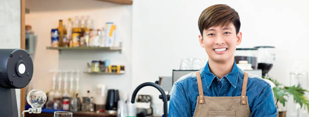 Cheerful young Asian man entrepreneur in coffee shop Cheerful young Asian man entrepreneur standing at counter in his own coffee shop, panoramic banner bar drink establishment photos stock pictures, royalty-free photos & images