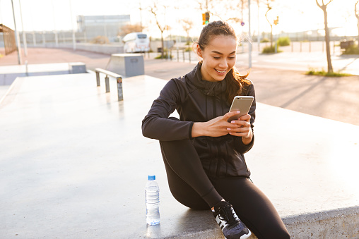 Photo of cheerful young asian sports woman sitting outdoors using mobile phone.