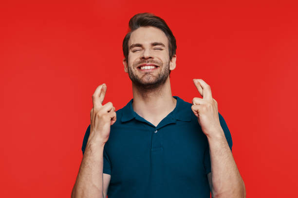 Cheerful young man in casual wear keeping fingers crossed stock photo