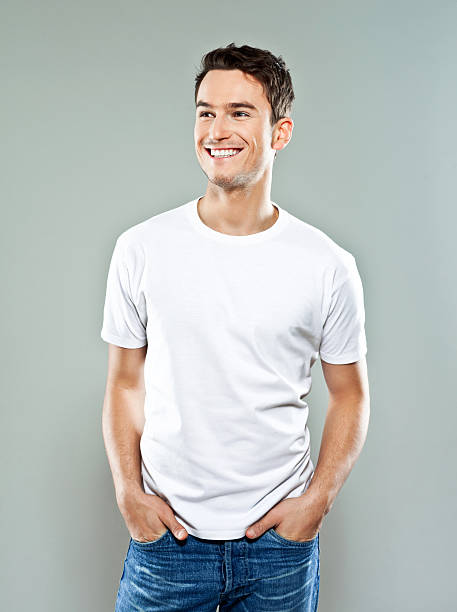 Cheerful young man Portrait of happy young man wearing white t-shirt, standing with hands in pockets and smiling. Studio shot, grey background. white t shirt stock pictures, royalty-free photos & images