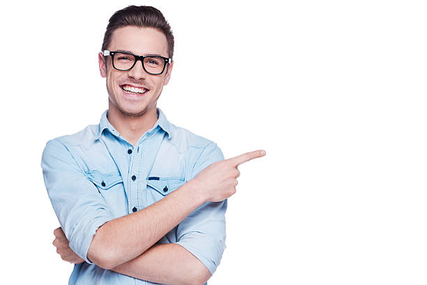 Cheerful young man pointing copy space. Handsome young man in shirt looking at camera and pointing away while standing against white background one young man only stock pictures, royalty-free photos & images