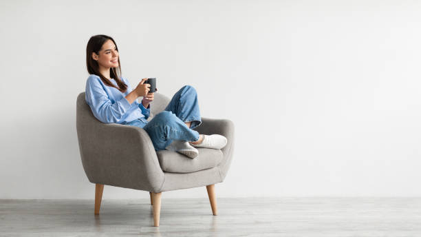 Cheerful young woman drinking aromatic coffee while chilling in comfy armchair against white wall, panorama Cheerful young woman drinking aromatic coffee while chilling in comfy armchair against white wall, panorama with blank space. Smiling millennial lady having hot beverage, relaxing during her break hot arab woman stock pictures, royalty-free photos & images