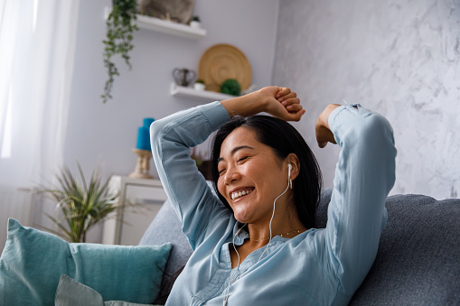 Candid shot of cheerful young woman sitting on the sofa with closed eyes, hands above her head, dancing and having fun listening to her favorite song via headphones.