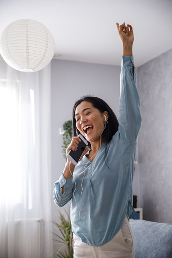 Portrait of cheerful young woman standing with arm raised and singing into her smart phone, as if it is a microphone, while listening to music via headphones.