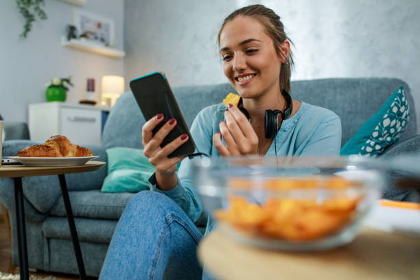 Cheerful young woman snacking on tortilla chips and using smart phone stock photo