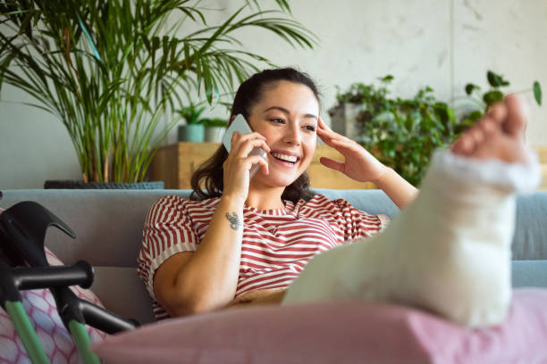 Cheerful young woman with broken leg on phone stock photo