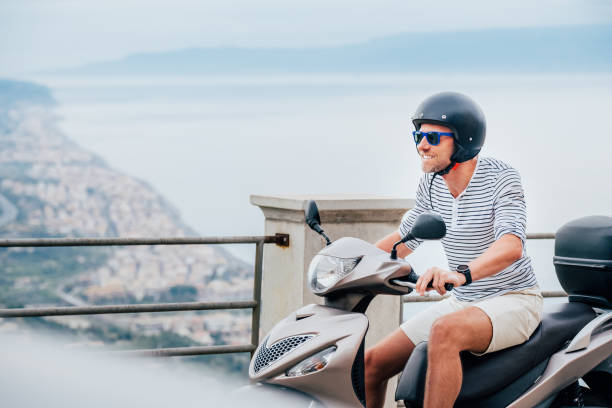 Cheerfully smiling man in helmet and sunglasses fast riding the moto scooter on the Sicilian old town streets on the Ionian Seaside coast. Happy Italian vacation and transportation concept. stock photo