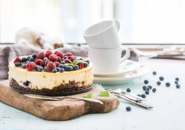 Cheesecake with fresh raspberries and blueberries on a wooden serving Cheesecake with fresh raspberries and blueberries on a wooden serving board, plates, cups, kitchen napkin, silverware over blue background, window at the backdrop tart dessert photos stock pictures, royalty-free photos & images