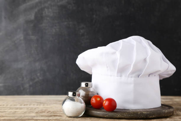 Chef hat with salt, pepper and tomato on wooden table stock photo