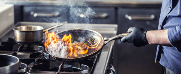 Chef in the kitchen leans pan to the side for alcohol in it to catch fire for gas stove, starting to flambe the food. stock photo