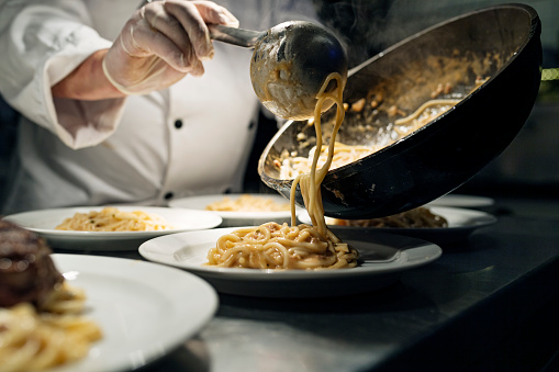 Chef pouring a serving of spaghetti with a tomato sauce from a saucepan to a plate at the pass. Photographed on location in a restaurant on the island of Møn in Denmark. Colour, horizontal with some copy space.