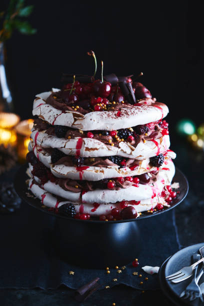 Cherry Chocolate Pavlova A five layer cherry chocolate pavlova with red currants, blackberries, cherries, chocolate curls, and golden sprinkles and stars with Christmas baubles and lights in the background pavlova dessert photos stock pictures, royalty-free photos & images