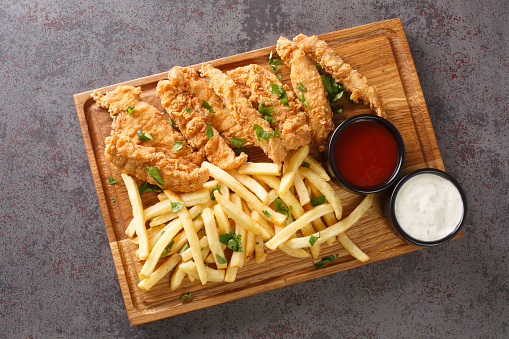 Chicken strips with ketchup, mayonnaise and french fries closeup on the wooden tray on a old concrete table. Horizontal top view from above