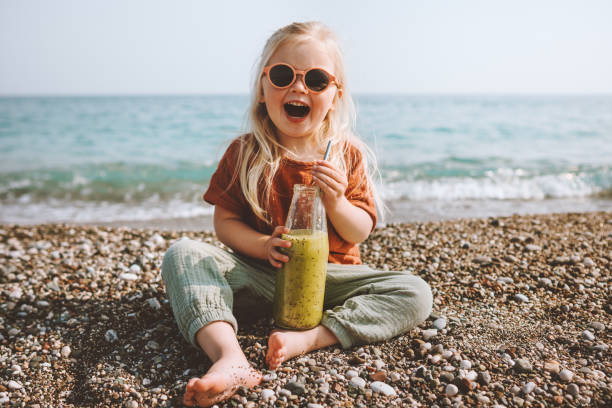 Child drinking smoothie on beach healthy lifestyle summer vacations child with reusable glass bottle vegan organic beverage picnic outdoor kid laughing happy emotions stock photo