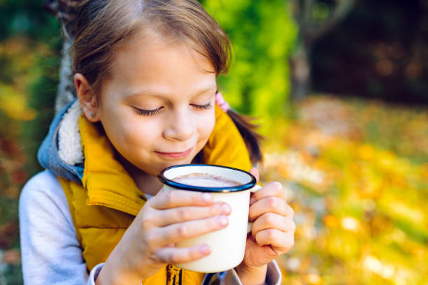 child girl drinking chocolate from a cup dressed in a warm yellow vest in autumn scenery stock photo